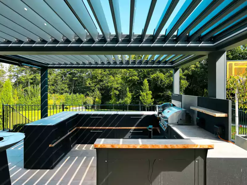 Outside kitchen under a pergola with a gas bbq, pizza oven, sink and bar overlooking a large garden.