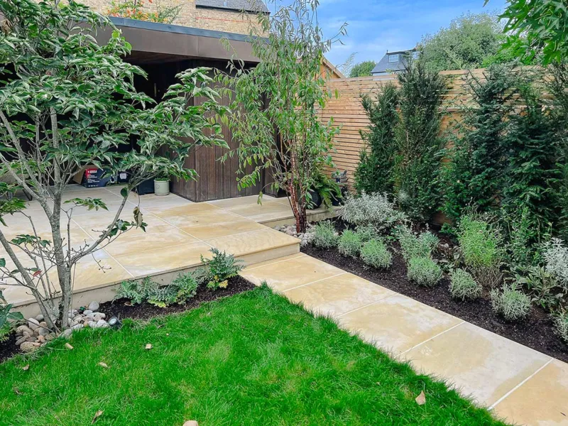 Medium garden with lots of rich green planting that is accentuated by a sandstone terrace and path. Behind the planting is a bespoke Venetian fence