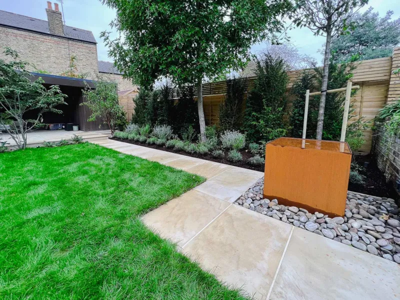 Sandstone pathway that runs beside a lawn with planting on one side with a yew hedge, lavender and small trees, including a liquidambar. Also, a corten water feature with a rusty effect.