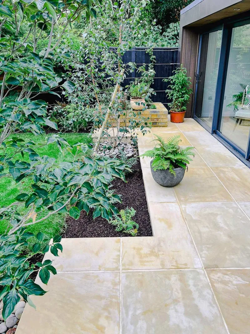 Small patio that is made from Smooth Harvest Sandstone that was constructed by luxury garden design in Teddington. London. There is a garden room to the right, with full glass frontage, and a lovely mixture of green planting. 