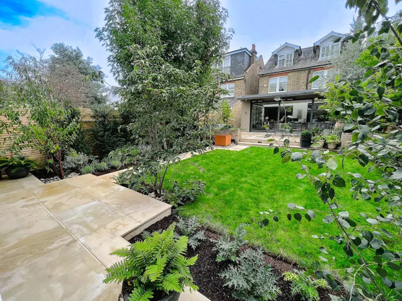 Medium sized garden with a small lawn space, sandstone paving, lots of planting and a corten water feature in the background.