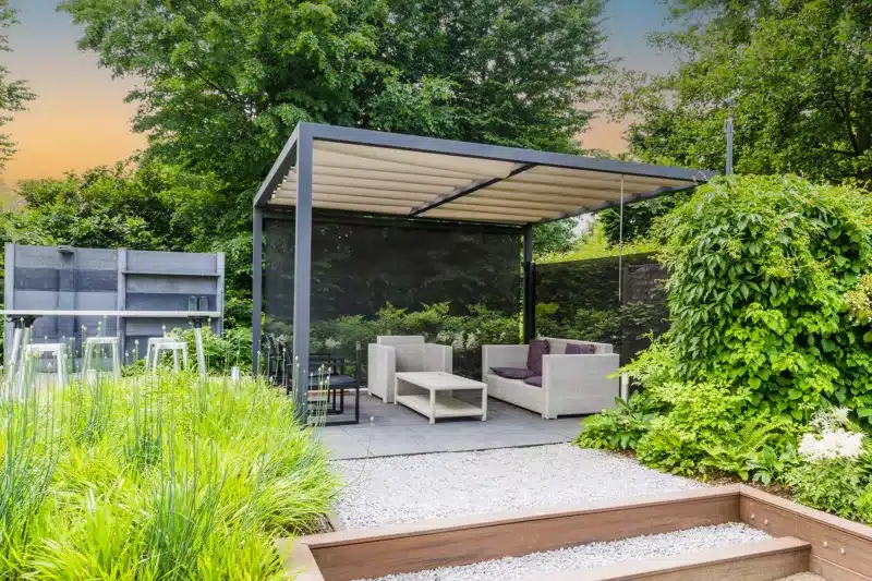 Contemporary garden with a simple pergola, gravel path, grey paving and modern kitchen in the background. The sun is setting with a blue and orange hue behind the trees.