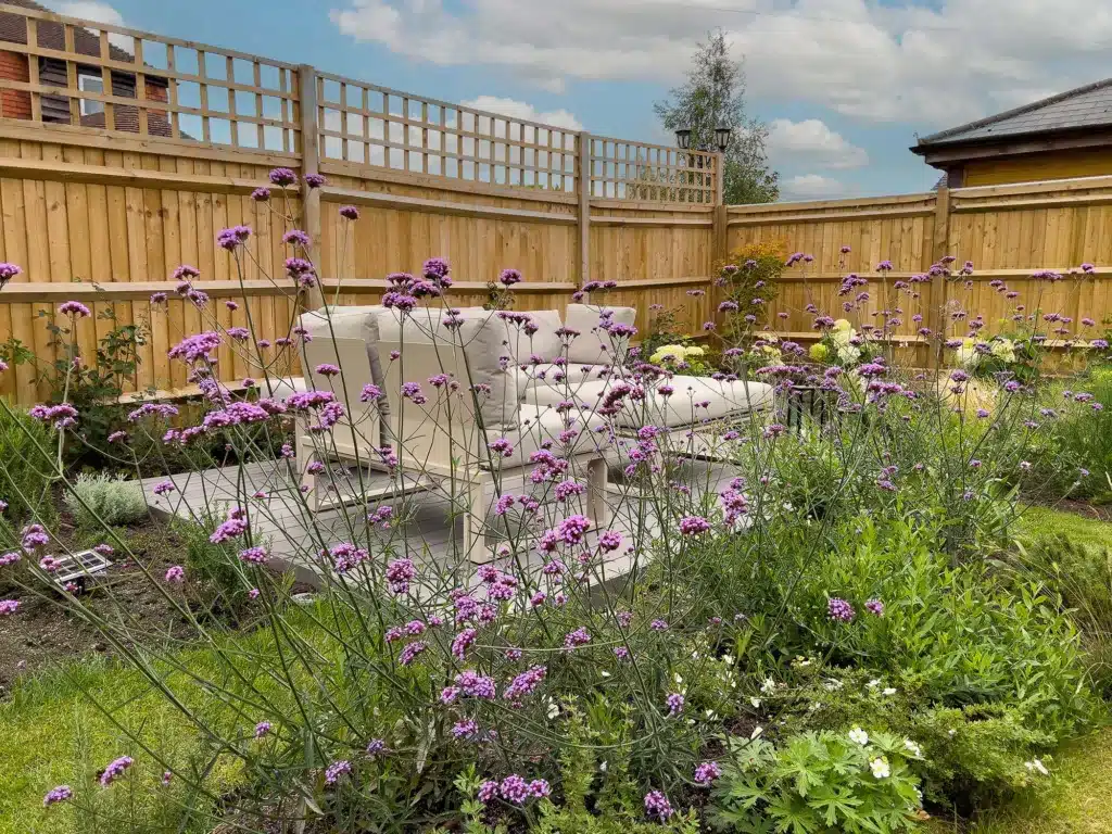 A garden full of colourful plants in the summer with verbena flowering in purple. Behind the flowers are a grey deck, wooden fence, and blue sky. This image is of a garden designed and built by Luxury Garden Design in West Sussex.